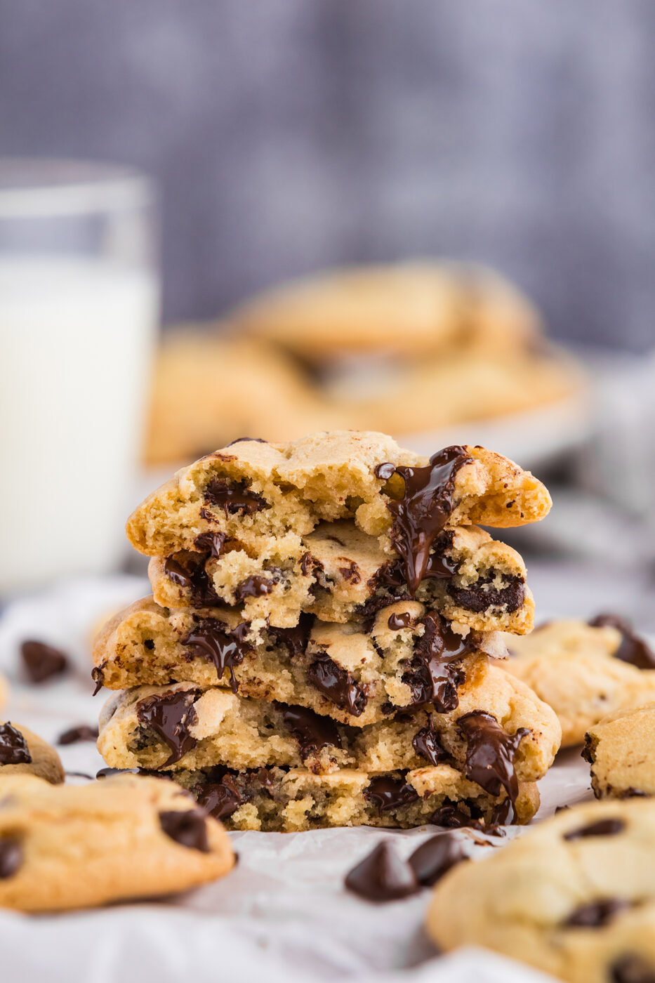 A stack of ooey, gooey broken chocolate cookies with the chocolate dripping down the front. More cookies and milk in the background