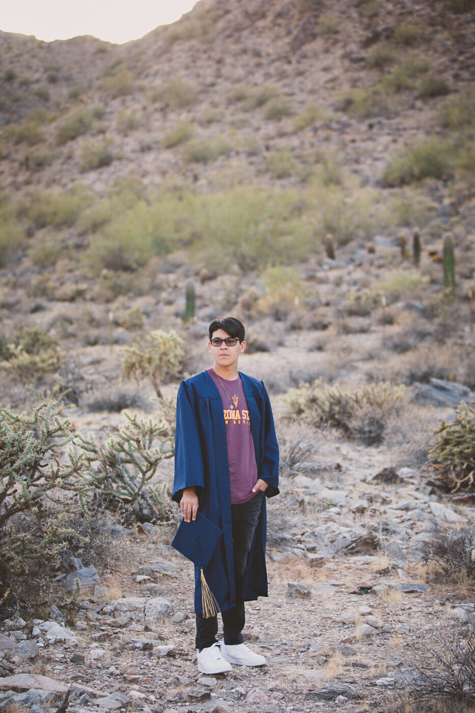 High school senior wearing an ASU shirt with a navy cap and gown on in the desert.