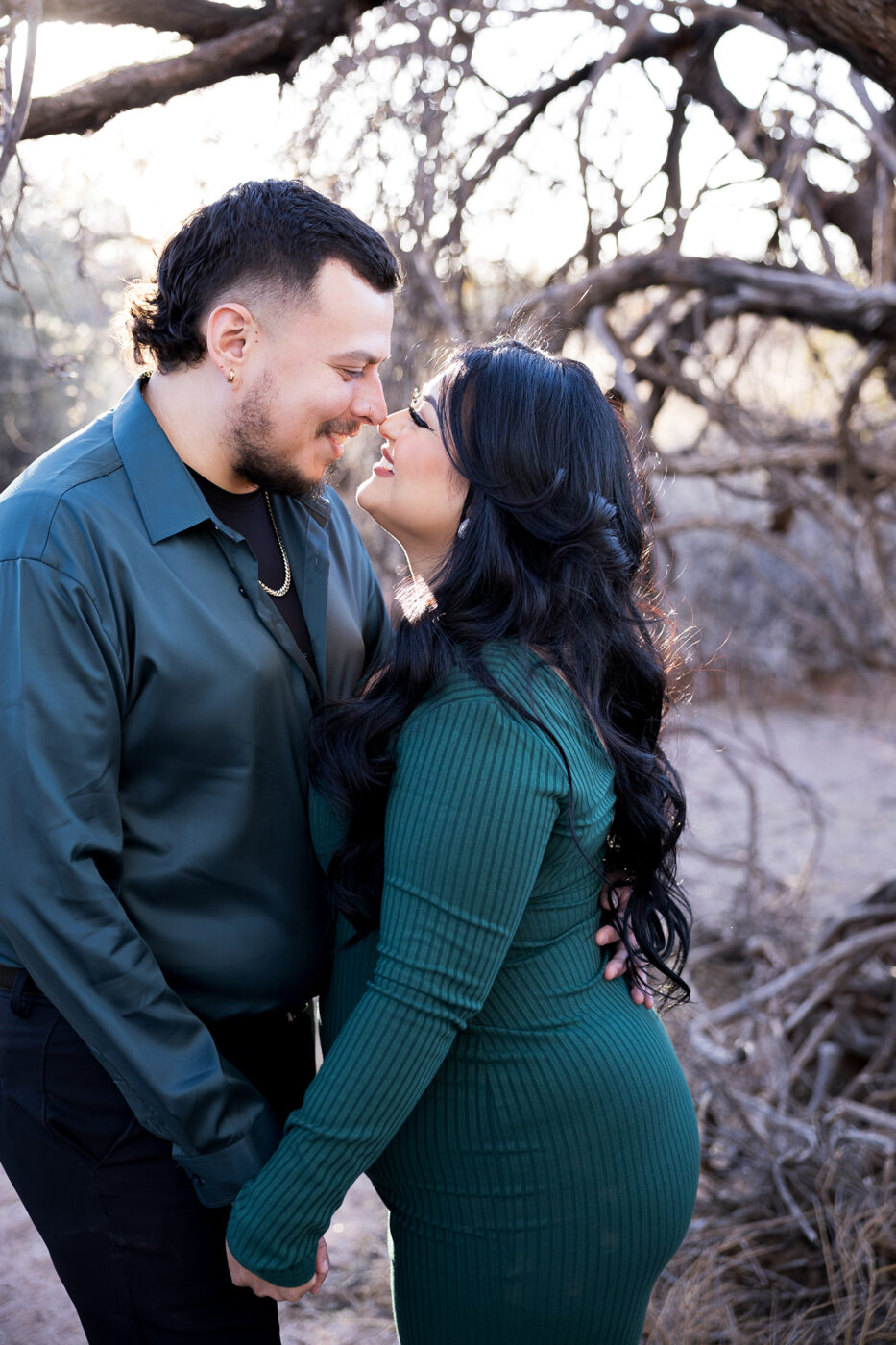 Engaged couple holding hands facing each other and smiling nose to nose with a craggly tree behind them.