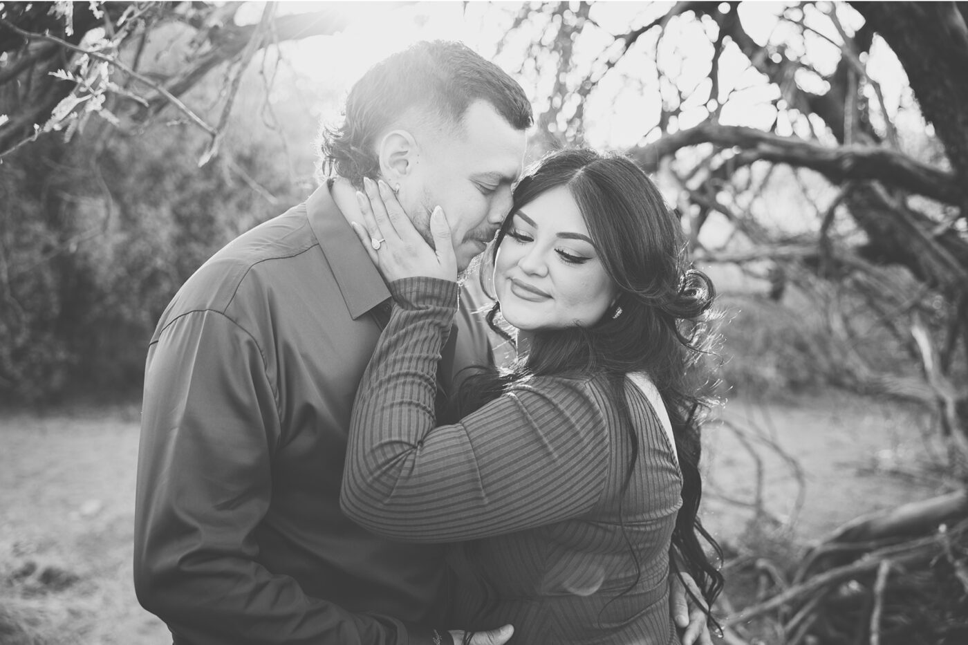 Engaged couple in desert trees, she's looking down holding his cheek and he's putting his nose in her hair.