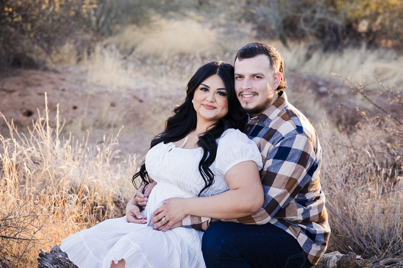 Couple sitting on a log in the desert with the woman cuddled up to him and both looking at the camera.