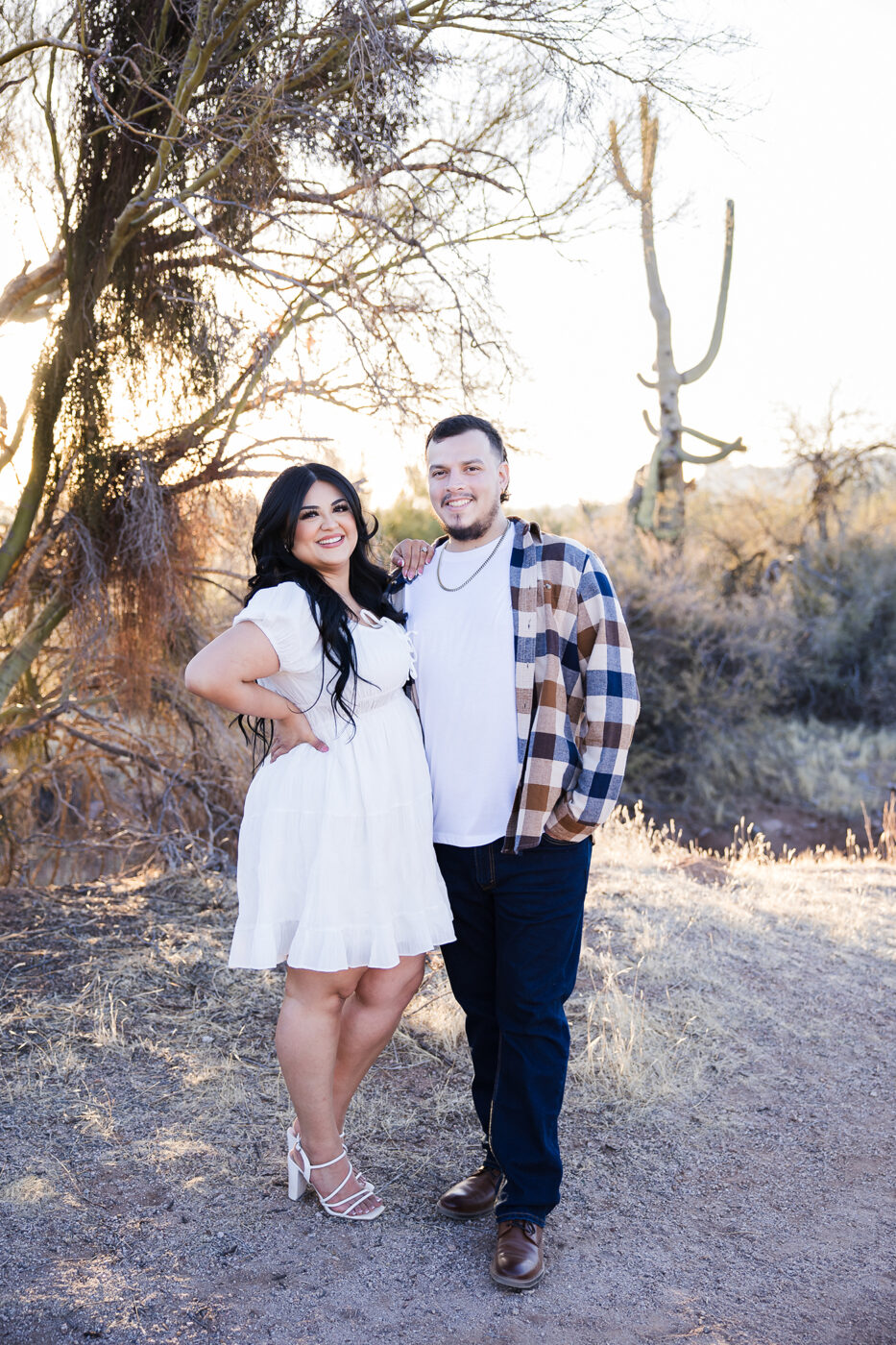 Full body image of an engaged couple in the desert, both are smiling.