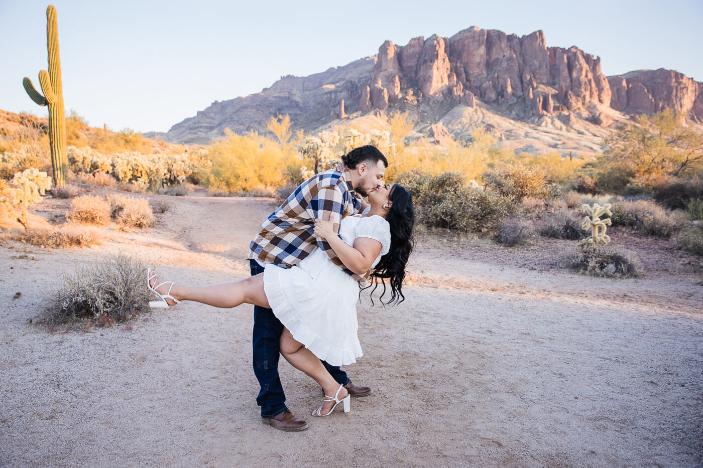 Full body shot of an engaged couple doing a dip kiss in front of Superstition Mountain at sunset.