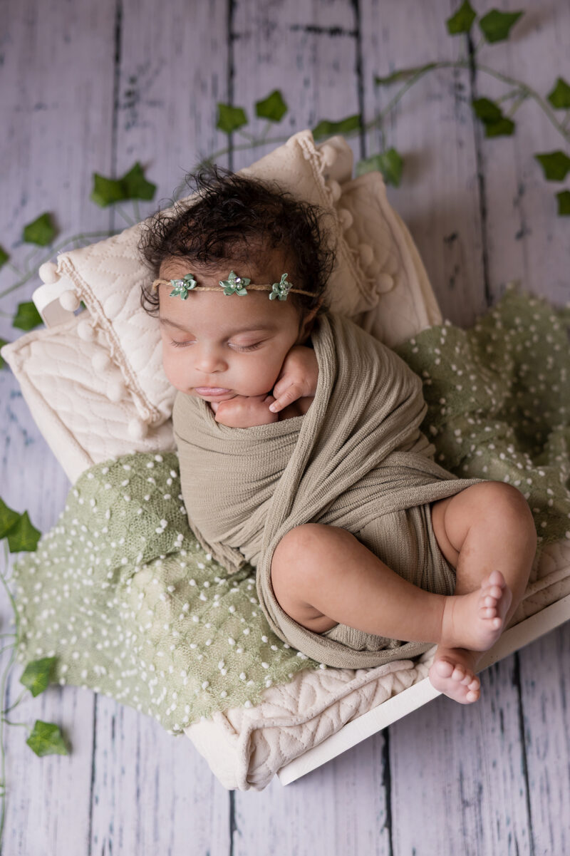 Sleeping newborn baby in a bed, wrapped in a pale green wrap with leaves on the white plank ground.