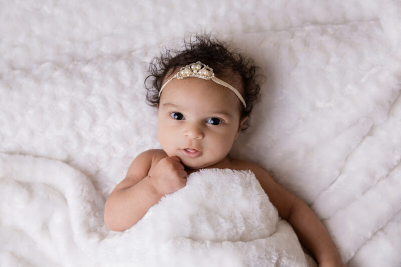Newborn baby girl with a crown on her head. Her eyes are open and in surrounding by white.