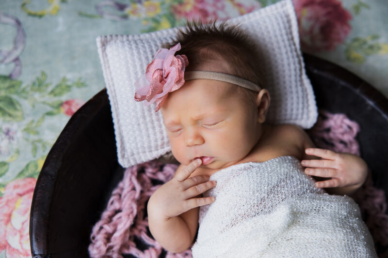 Sleeping newborn baby girl with a pink bow on her head. She's wrapped in cream and laying a a bowl on a floral background.