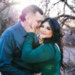 Engaged couple in desert trees with sun flare, she's looking at the camera holding his cheek and he's putting his nose in her hair.