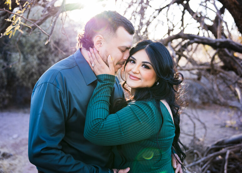 Engaged couple in desert trees with sun flare, she's looking at the camera holding his cheek and he's putting his nose in her hair.