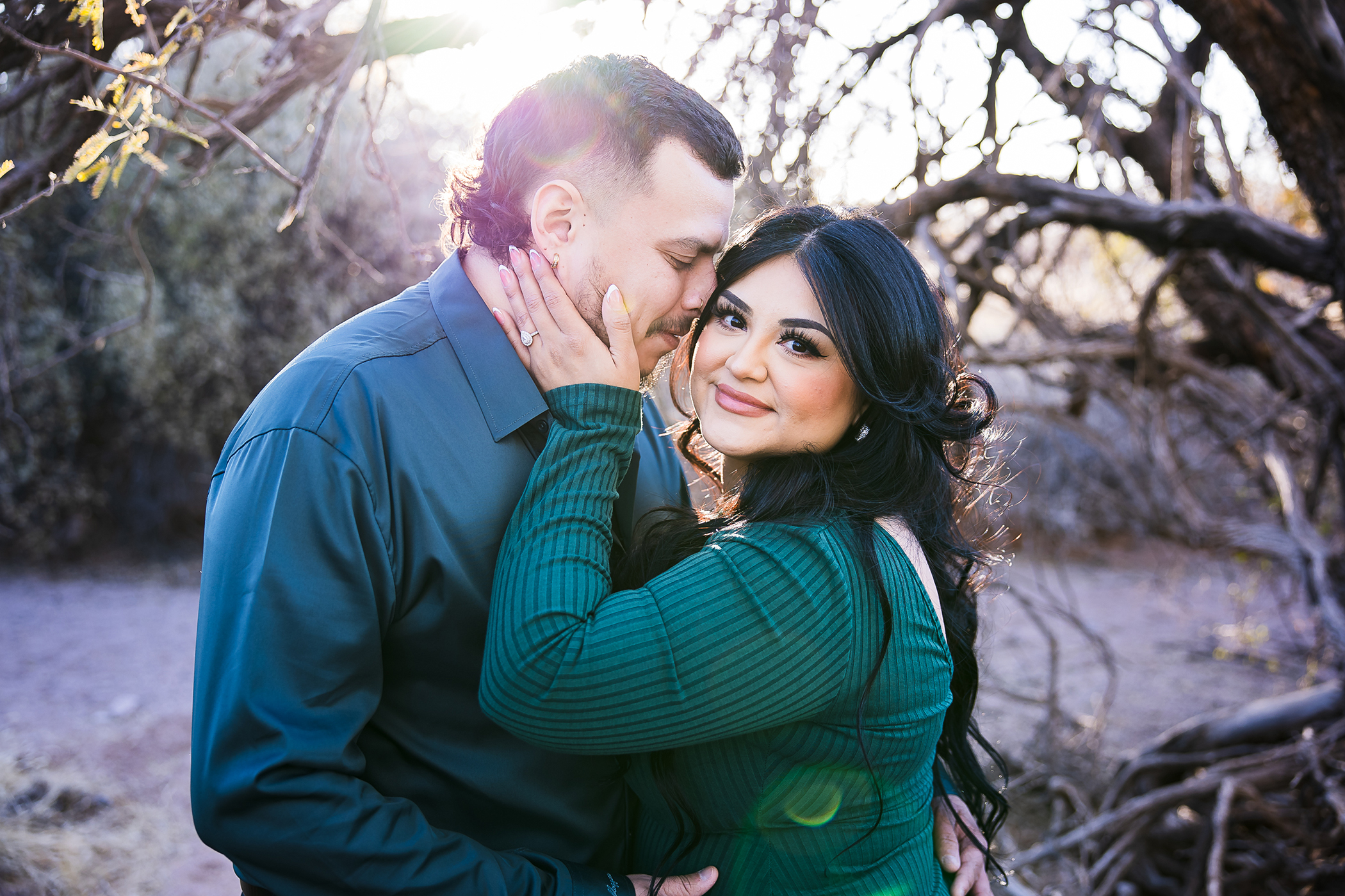 Engaged couple in desert trees with sun flare, she's looking at the camera holding his cheek and he's putting his nose in her hair.