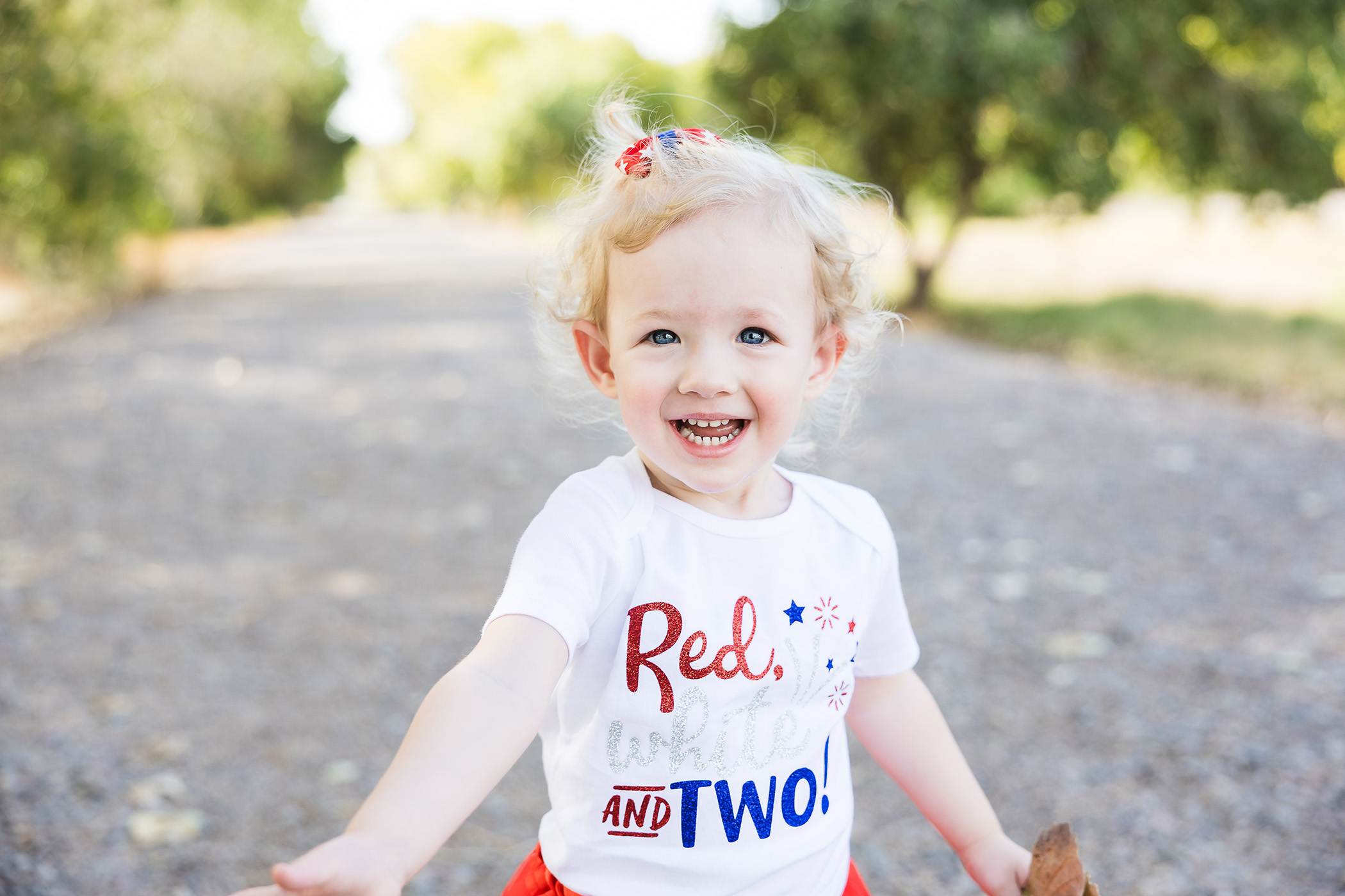 2 year old girl in patriotic clothes smiling in a tree area.
